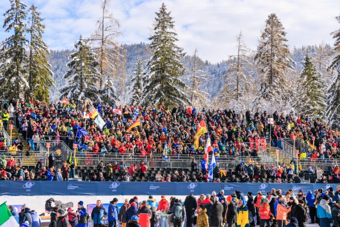 Große Zuschauertribüne mit Fans und Fahnen vor verschneiter Waldkulisse.