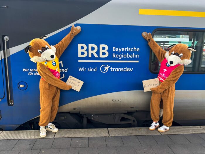 Zwei Beppo-Fuchs-Maskottchen stehen vor einem BRB-Zug und halten Schilder zum Biathlon-Weltcup 2026 in Ruhpolding hoch.